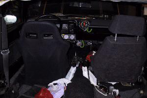 The stripped interior, with a pair of black bucket seats, harnesses, a roll cage bar and extra gauges on the dash