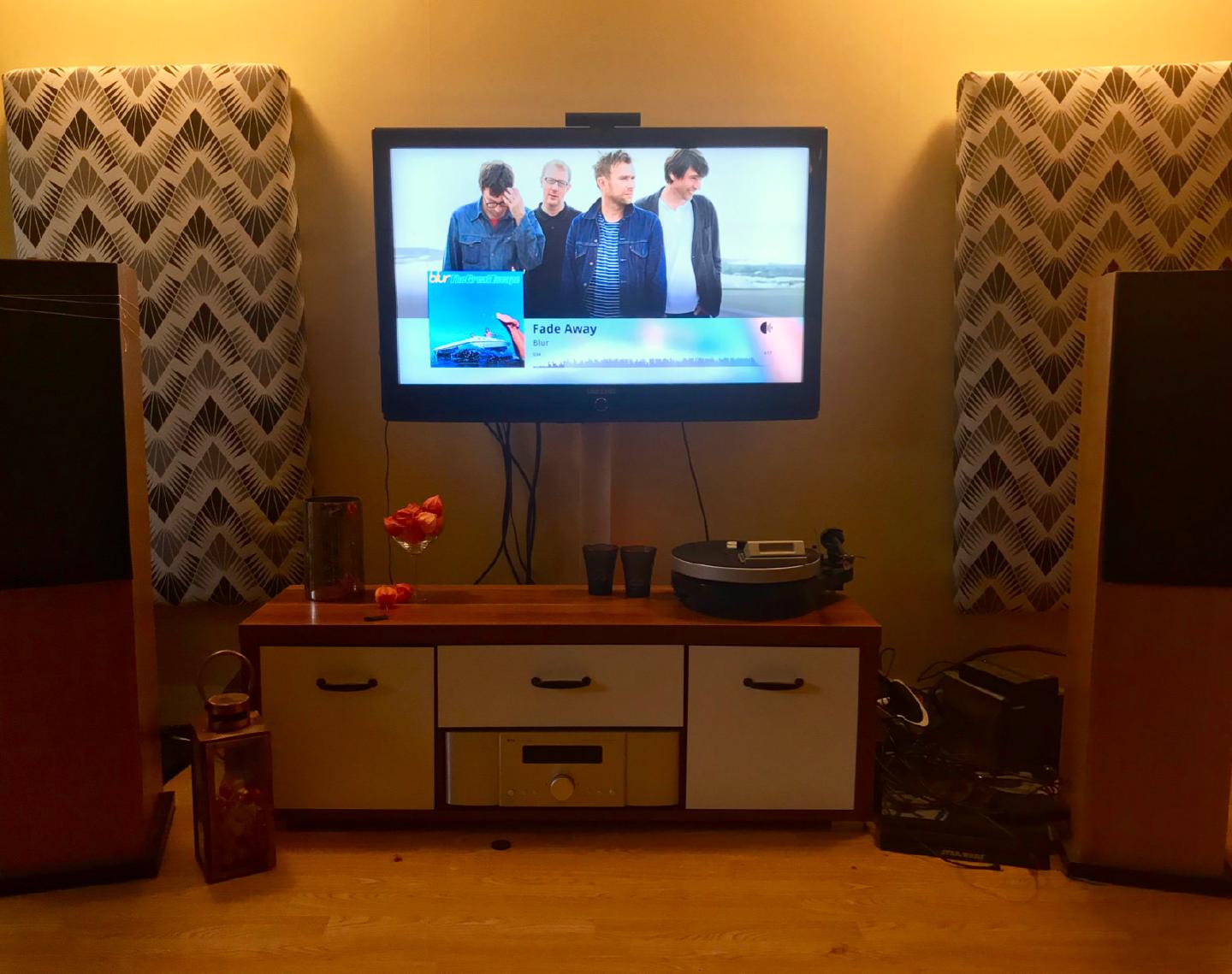 A wall-mounted TV running Roon’s display mode, showing artwork and a waveform for Blur’s Fade Away between two floorstanding speakers