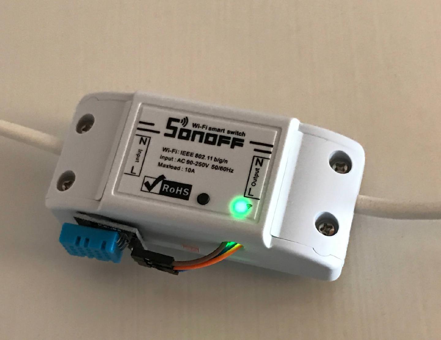 A wall-mounted Sonoff with its Wi-Fi LED lit green and a blue DHT11 temperature and humidity sensor hanging off the front on a short ribbon